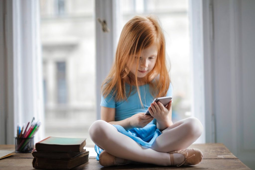 Red haired charming schoolgirl in blue dress browsing smartphone while sitting on rustic wooden table with legs crossed beside books against big window at home